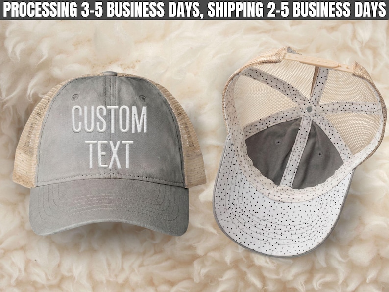 May include: A grey trucker hat with a mesh back and the words CUSTOM TEXT embroidered in white on the front. The interior of the hat features a white lining with a black polka dot pattern, and an adjustable snapback closure is visible at the back.