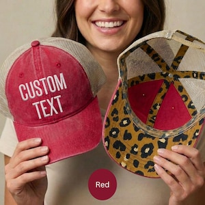 May include: Red trucker hat with "CUSTOM TEXT" embroidered in white. The hat has a mesh back and a red front. The underside of the brim features a leopard print design. The hat is a stylish accessory.