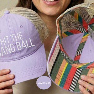 May include: A lavender trucker hat with a tan mesh back and the white text HIT THE DANG BALL on the front. The interior of the hat features a colorful striped pattern in red, yellow, green, and black along the seams and the underside of the brim.