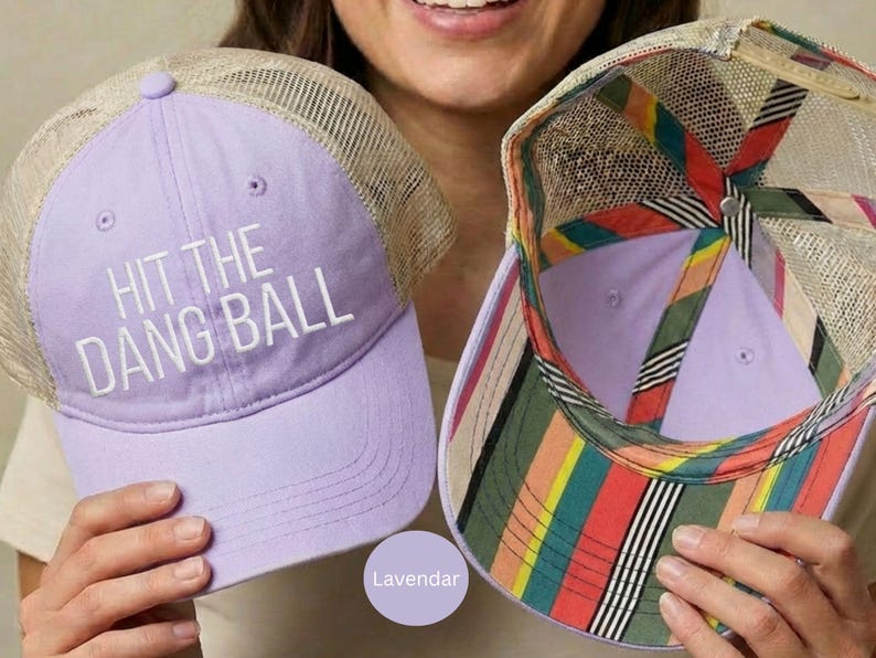 May include: A lavender trucker hat with a tan mesh back and the white text HIT THE DANG BALL on the front. The interior of the hat features a colorful striped pattern in red, yellow, green, and black along the seams and the underside of the brim.