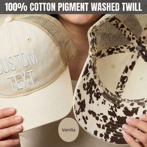 May include: A vanilla-colored trucker hat made of 100% cotton pigment washed twill. The front panel features the embroidered text CUSTOM TEXT in white. The hat has a mesh back and an interior lining with a brown and white cow print pattern.