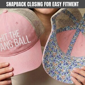 May include: A pink and beige mesh trucker hat featuring the white embroidered text HIT THE DANG BALL on the front panel. The interior of the hat displays a blue, pink, and green floral pattern along the seams and the underside of the brim.