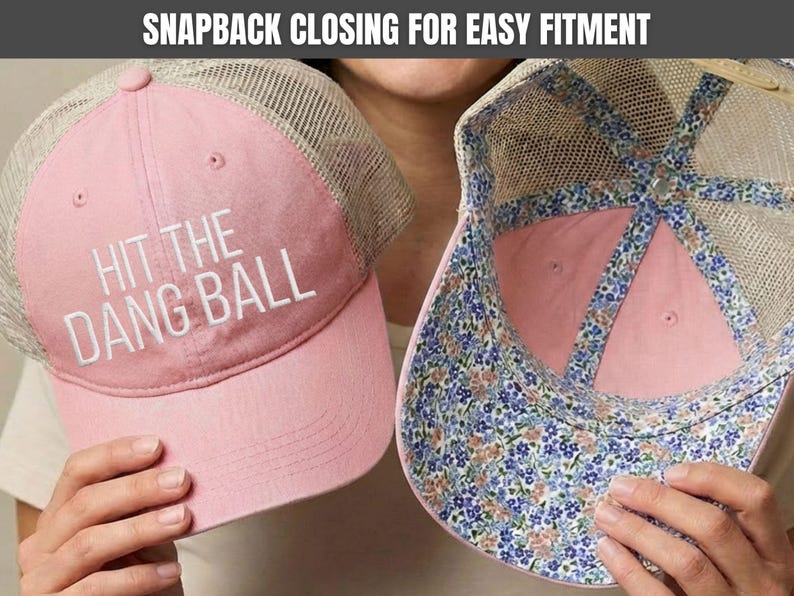 May include: A pink and beige mesh trucker hat featuring the white embroidered text HIT THE DANG BALL on the front panel. The interior of the hat displays a blue, pink, and green floral pattern along the seams and the underside of the brim.