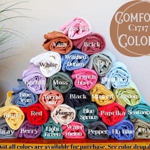 May include: A pyramid of rolled Comfort Colors C1717 t-shirts in various shades including blossom, yam, brick, white, washed denim, bay, orchid, moss, crunchberry, chambray, blue jean, terracotta, black, mustard, celedon, butter, red, ivory, blue spruce, paprika, seafoam, gray, berry, light green, watermelon, pepper, flo blue, and khaki.