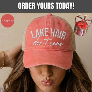 May include: A coral-colored baseball cap with a mesh back and the white embroidered text "LAKE HAIR don't care" on the front. The cap features a high ponytail opening at the back with a colorful striped pattern on the interior strap structure.