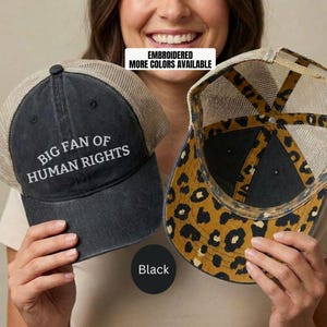 May include: Two black trucker hats with mesh backs. One hat has the text "BIG FAN OF HUMAN RIGHTS" embroidered in white. The other hat has a leopard print design on the underside. The text "EMBROIDERED MORE COLORS AVAILABLE" is in a white rectangle.