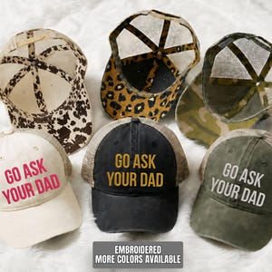 May include: A collection of six trucker hats with mesh backs. The hats feature various patterns, including animal print and camouflage. Three hats have the phrase "GO ASK YOUR DAD" embroidered on the front in different colors. The text "EMBROIDERED MORE COLORS AVAILABLE" is also visible.