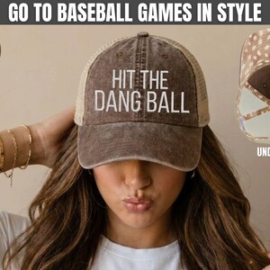 May include: A chocolate brown baseball cap with a mesh back and the white embroidered text HIT THE DANG BALL on the front. The hat features a distressed finish and a light brown interior lining with a white polka dot pattern visible in the inset.