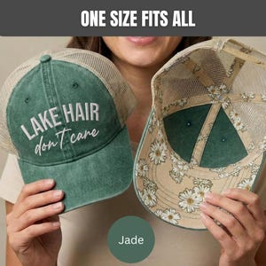 May include: A green trucker hat with a mesh back and the text "LAKE HAIR don't care" embroidered in white on the front. The interior of the hat features a white daisy floral pattern on a tan background. The text "ONE SIZE FITS ALL" is visible.