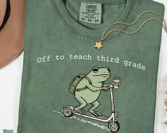 May include: A moss green crew neck t-shirt featuring a graphic of a frog wearing a backpack and riding a scooter while holding a coffee cup. The text "Off to teach third grade" is printed in white above the frog. The shirt has a Comfort Colors label.