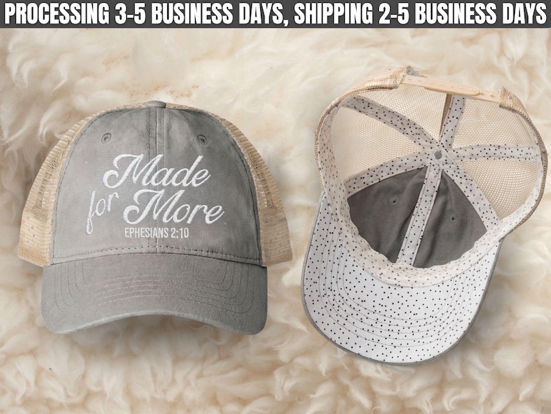 May include: A grey trucker hat with a tan mesh back and an adjustable strap. The front features white embroidered text that reads Made for More EPHESIANS 2:10. The interior lining of the cap displays a white fabric with a black polka dot pattern.