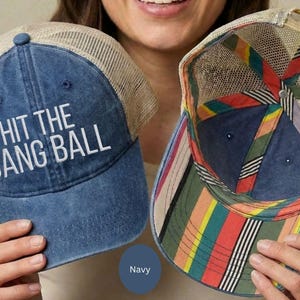 May include: A navy blue baseball cap with a tan mesh back and the white embroidered text HIT THE DANG BALL on the front. The interior of the cap features a colorful striped pattern in red, yellow, green, and black along the inner seams.