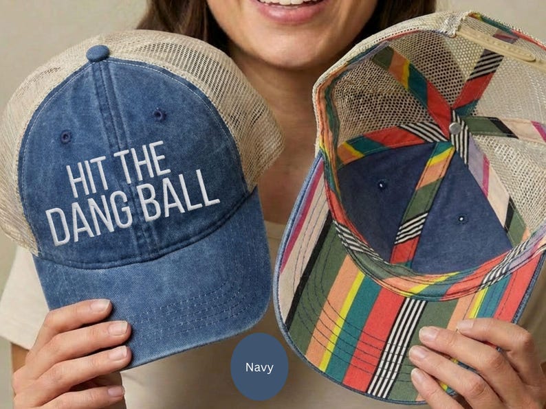 May include: A navy blue baseball cap with a tan mesh back and the white embroidered text HIT THE DANG BALL on the front. The interior of the cap features a colorful striped pattern in red, yellow, green, and black along the inner seams.