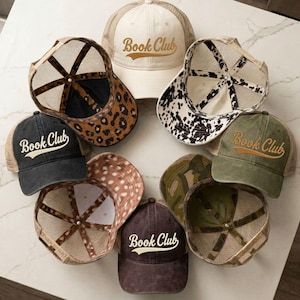 May include: A collection of seven trucker hats arranged in a circle, each featuring the text Book Club embroidered on the front. The hats include various patterns such as leopard print, cow print, camouflage, and solid colors with mesh backs.