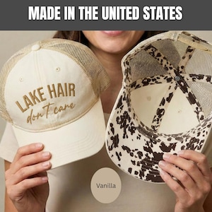May include: Two trucker hats with mesh backs. One hat is cream-colored with the embroidered brown text 'LAKE HAIR don't care' on the front. The other hat shows the interior, featuring a brown and white cow print pattern on the underside of the brim.