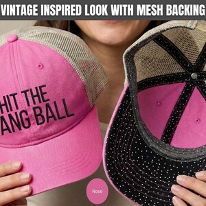 May include: A bright pink baseball cap with a beige mesh back and the text HIT THE DANG BALL embroidered in black on the front. The interior of the cap features a black lining with white polka dots and a pink crown, with a vintage inspired look.