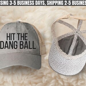 May include: A gray trucker hat with a tan mesh back and a curved brim. The front panel features the black text HIT THE DANG BALL. The interior of the hat displays a white lining with a black polka dot pattern and a tan adjustable snapback closure.