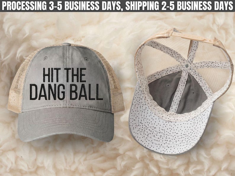 May include: A gray trucker hat with a tan mesh back and a curved brim. The front panel features the black text HIT THE DANG BALL. The interior of the hat displays a white lining with a black polka dot pattern and a tan adjustable snapback closure.