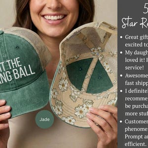 May include: A green trucker hat with a mesh back and the white text HIT THE DANG BALL on the front. The interior of the hat features a beige lining with a white daisy floral pattern. The hat has a structured front panel and a curved brim.