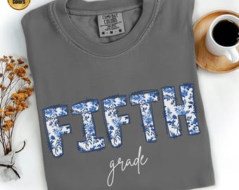 Könnte beinhalten: Ein graues Comfort Colors T-Shirt mit dem Wort "FIFTH" in großen blauen Blumenbuchstaben und dem Wort "grade" in Kursivschrift. Eine Kaffeetasse und ein kleiner Strauß getrockneter Blumen befinden sich rechts und links vom T-Shirt.