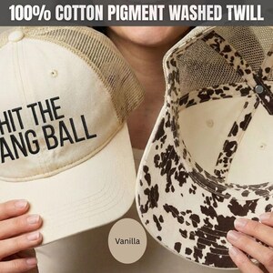 May include: A vanilla-colored trucker hat made of 100% cotton pigment washed twill. The front panel features the black text HIT THE DANG BALL. The hat has a mesh back and an interior lining with a brown and white cow print pattern.