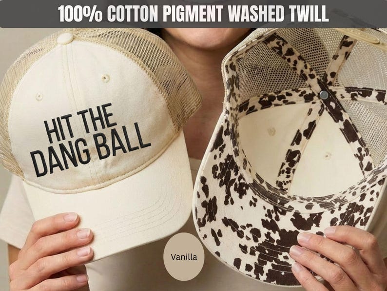 May include: A vanilla-colored trucker hat made of 100% cotton pigment washed twill. The front panel features the black text HIT THE DANG BALL. The hat has a mesh back and an interior lining with a brown and white cow print pattern.