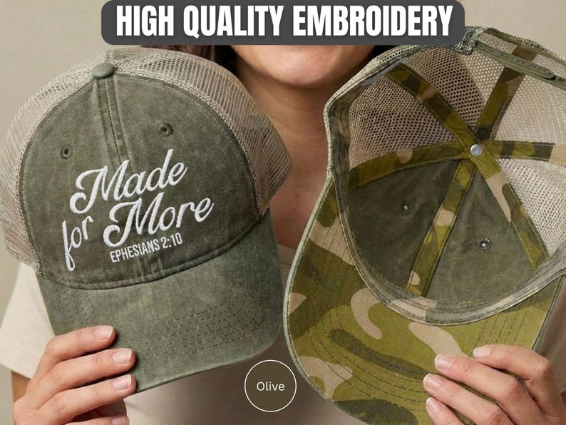 May include: An olive green trucker hat with a mesh back and white embroidered text that reads Made for More EPHESIANS 2:10. The interior of the hat features a camouflage pattern on the underside of the brim and along the inner structural seams.