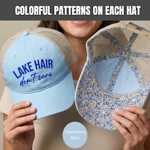 May include: Two light blue trucker hats with beige mesh backs. One hat features the dark blue text "LAKE HAIR don't care" on the front. The other hat is shown from the inside, revealing a floral pattern lining with blue and orange flowers.