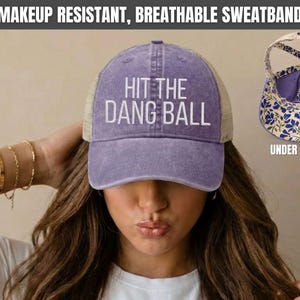 May include: A violet baseball cap with a mesh back and the white text HIT THE DANG BALL embroidered on the front. The hat features a makeup resistant, breathable sweatband and an interior lining with a blue and white floral pattern.