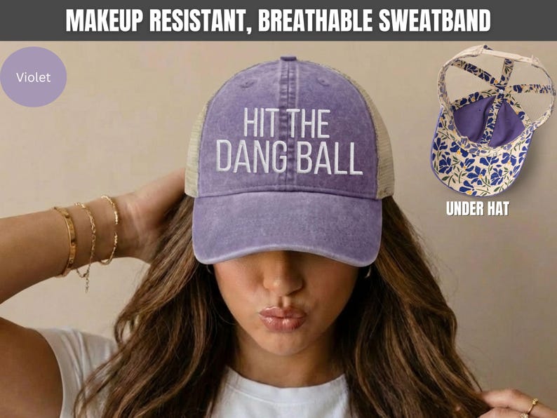 May include: A violet baseball cap with a mesh back and the white text HIT THE DANG BALL embroidered on the front. The hat features a makeup resistant, breathable sweatband and an interior lining with a blue and white floral pattern.