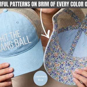 May include: A light blue trucker hat with a mesh back and the white embroidered text HIT THE DANG BALL on the front. The interior of the brim and the inner seams feature a colorful floral pattern with small blue and peach flowers on a white base.