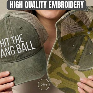 May include: An olive green trucker hat with a mesh back and the white embroidered text HIT THE DANG BALL on the front. The interior of the hat features a camouflage pattern on the underside of the brim and along the inner crown panels.
