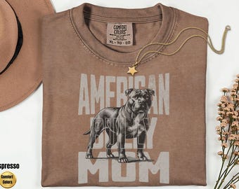 May include: Espresso-colored t-shirt with the text "AMERICAN BULLY MOM" and a graphic of a pit bull. A gold star pendant necklace and a brown hat are visible. The shirt is from Comfort Colors.