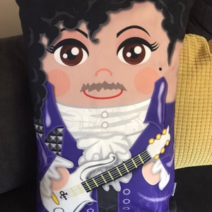 Może przedstawiać: Poduszka w kolorze fioletowym i białym z kreskówkowym wizerunkiem Prince'a grającego na gitarze. Poduszka ma czarne tło i białą obwódkę.