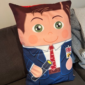 May include: A cushion with a cartoon illustration of a man in a blue suit and red tie, holding a flashlight. The pillow has a light skin tone and green eyes. The FBI logo is visible on the cushion.