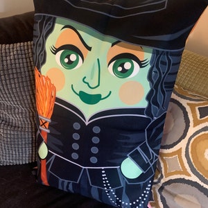 Peut inclure: Un coussin de sorcière noir et vert avec un chapeau noir, un balai orange et un collier de perles blanches. La sorcière a un visage vert avec de grands yeux et une bouche noire.