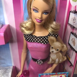 May include: A blonde Barbie doll wearing a pink dress with black polka dots and a black belt with a silver buckle. She is holding a pink purse and has a brown and white puppy dog sitting on her arm. The doll is standing in front of a pink and white striped background with a sign that says "Barbie Doll for a Day! Instant win sweepstakes!"