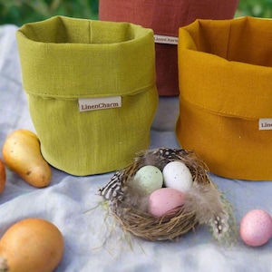 Puede incluir: Tres cestas de almacenamiento de tela en diferentes colores: verde, naranja y marrón. Las cestas están hechas de lino y tienen una etiqueta que dice "LinenCharm".