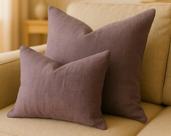 May include: Two decorative throw pillows in a deep purple hue, made of a soft, textured fabric. The pillows are arranged on a light beige couch.