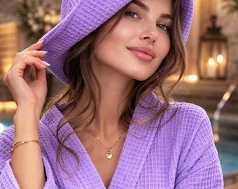 May include: A woman wearing a lavender waffle-weave robe and matching hooded towel. The robe has a shawl collar and is secured with a belt. The hooded towel covers the head. The background is blurred, suggesting a spa or relaxation setting.