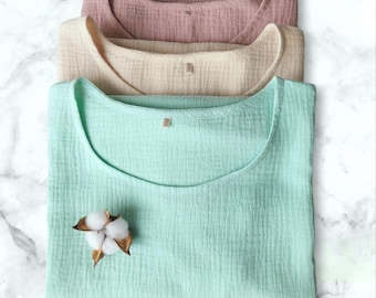 May include: Three cotton muslin tops in different colors: light green, beige, and dusty rose. The tops are folded and stacked on a white marble surface. A small cotton flower is placed on the top of the green top.