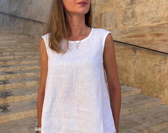 May include: A white linen sleeveless top and matching trousers. The outfit is accessorised with sunglasses and a silver necklace. The background features a stone wall and steps, suggesting an outdoor setting. The overall style is casual and summery.