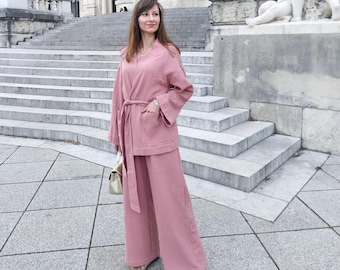 May include: A woman wearing a blush pink wide-leg pantsuit with a matching long-sleeved jacket. The jacket has a tie belt and pockets. She is wearing gold sandals and carrying a small gold handbag.