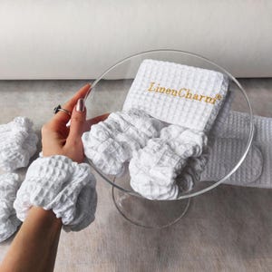 May include: Set of white waffle weave fabric hair scrunchies and a headband with the text "Linen Charm" embroidered in gold thread.