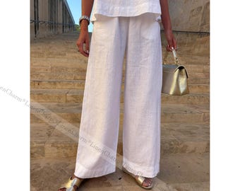 May include: White linen wide-leg pants paired with a matching top. The outfit is accessorized with gold sandals and a gold handbag. The model is standing on stone steps, creating a casual, yet elegant look.