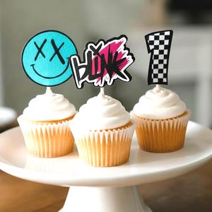 May include: Three cupcakes topped with white frosting and decorative toppers. One topper is a teal smiley face with X eyes, one says BLINK in black and pink, and one is a black and white checkered number one. The cupcakes sit on a white stand.