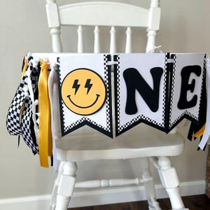 May include: A white high chair decorated with a banner that says "ONE". The banner has black and white checkered flags, yellow ribbons, and a yellow smiley face with lightning bolts. The letters are black.