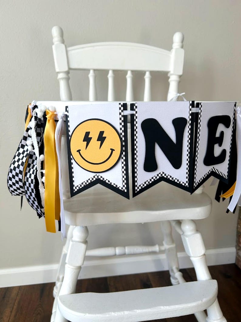 May include: A white high chair decorated with a banner that says "ONE". The banner has black and white checkered flags, yellow ribbons, and a yellow smiley face with lightning bolts. The letters are black.
