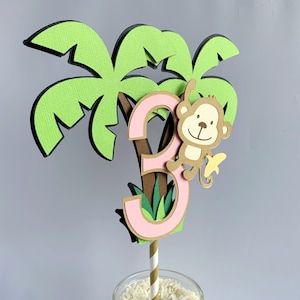 May include: A pink number three cake topper with a brown monkey holding a yellow banana. The number is surrounded by green palm leaves and a brown palm tree trunk.