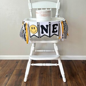 May include: A white high chair decorated for a first birthday. The chair has a banner that says "ONE" with a smiley face and lightning bolt graphic. The banner is black and white with yellow accents. A small cake sits on the tray.